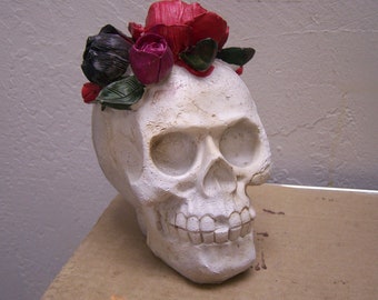 May include: White ceramic skull with a flower crown made of red, pink, and black fabric flowers.