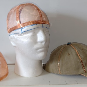 May include: A selection of baseball caps, including a finished olive green cap with copper-coloured accents. Also shown are two transparent hat forms with copper-coloured tape, one on a mannequin head.