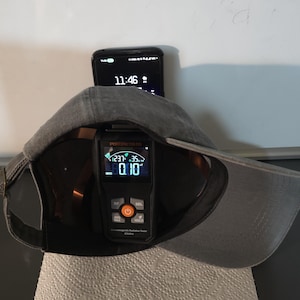 May include: A gray baseball cap with a black digital radiation tester inside. The tester displays readings and has orange and gray buttons. A smartphone showing 11:46 is behind the cap.
