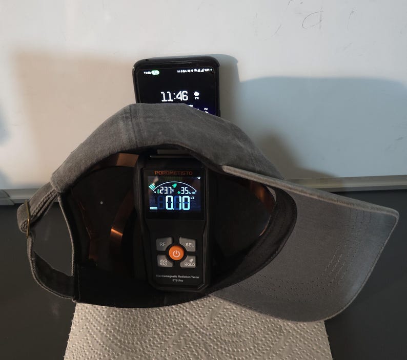 May include: A gray baseball cap with a black digital radiation tester inside. The tester displays readings and has orange and gray buttons. A smartphone showing 11:46 is behind the cap.