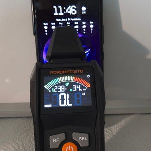 May include: Black electromagnetic radiation tester with a digital display showing readings. The device has buttons labeled RF, SEL, AVG MAX, and HOLD. The screen displays the text "POROMETISTO" and "Electromagnetic Radiation Tester ET01Pro". A smartphone is behind the device.