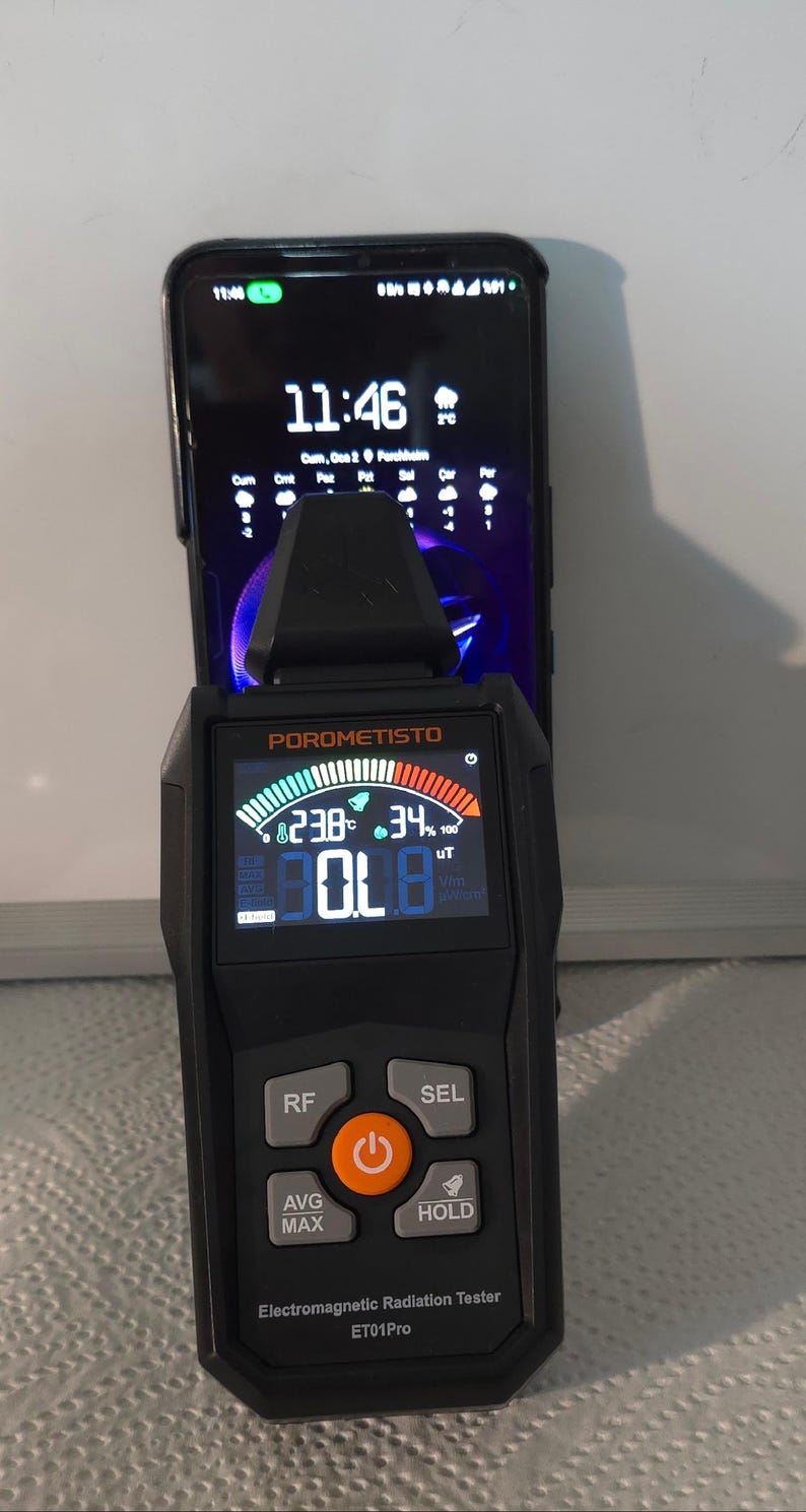 May include: Black electromagnetic radiation tester with a digital display showing readings. The device has buttons labeled RF, SEL, AVG MAX, and HOLD. The screen displays the text "POROMETISTO" and "Electromagnetic Radiation Tester ET01Pro". A smartphone is behind the device.