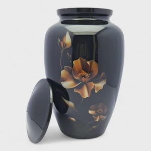 Op de afbeelding: Een glanzend zwarte urn met een afgerond lichaam en een bijpassend afneembaar deksel. Het oppervlak is voorzien van een geschilderd ontwerp van twee goudbruine bloemen met fijne stengels en bladeren, die verticaal over de voorzijde lopen.