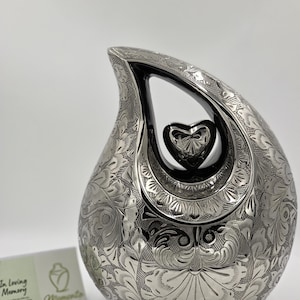 Puede incluir: Urna plateada con forma de lágrima y grabados florales intrincados. El diseño incluye una abertura en forma de corazón y un corazón más pequeño en el interior. Se ven las palabras "In Loving Memory" y "Memento Urns Ltd".