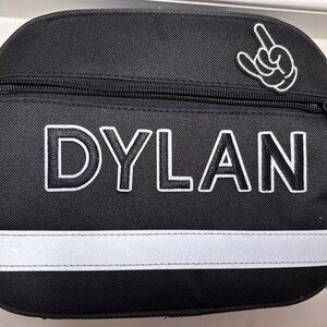 May include: Black lunch bag with the name "DYLAN" in white block letters. A white and black hand gesture patch is on the upper right. A white reflective stripe runs across the bottom. The bag has a black zipper.
