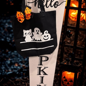 Puede incluir: Bolso tote negro de Halloween con un diseño de gato, fantasma y calabaza blancos. El bolso tiene un asa negra y un lazo de lunares naranja y amarillo. Un letrero de madera dice "hello" y "PKIN".