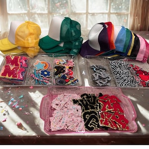Puede incluir: Una colección de gorras de camionero amarillas, verdes y moradas con frentes blancos, junto con bandejas de parches y apliques decorativos. Los parches incluyen letras, estrellas, flores y otros diseños, perfectos para personalizar ropa y accesorios.