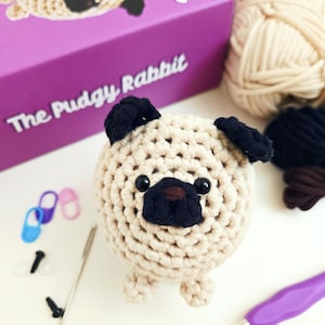 May include: A crocheted pug dog with black ears, nose, and eyes. The pug is beige and sits on a white surface. A purple box with the text "The Pudgy Rabbit" is in the background, along with yarn, stitch markers, and crochet hooks.
