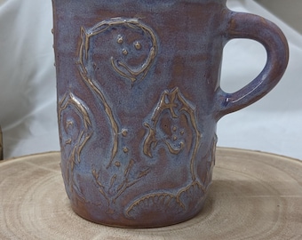 May include: A handmade ceramic mug with a unique design. The mug features a deep purple glaze with blue highlights and intricate embossed floral patterns. The handle is curved, and the mug sits on a wooden surface. A decorative piece for the kitchen.