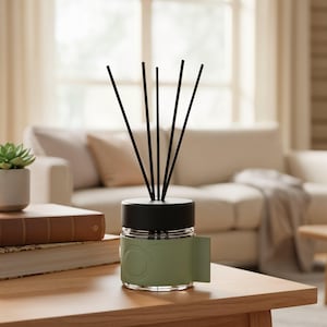 Puede incluir: Un difusor de caña con cañas negras, una tapa negra y un tarro de cristal transparente con una envoltura verde. El difusor se encuentra sobre una mesa de madera con libros y una pequeña planta en maceta. El fondo incluye un sofá y una ventana.