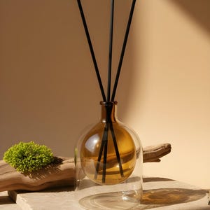 Könnte beinhalten: Bernsteinfarbener Glasdiffusor mit schwarzen Stäbchen. Der Diffusor hat eine kugelförmige Form in einer klaren Glasbasis. Die Stäbchen werden in den bernsteinfarbenen Glasteil gesteckt. Der Hintergrund ist neutral beige.