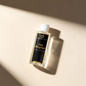 Könnte beinhalten: Eine transparente Glasflasche mit Oud Bergamot-Duft von MANGO.TYLE, mit weißem Verschluss und schwarzem Etikett. Das Etikett zeigt goldfarbene Schrift und ein goldenes kreisförmiges Logo. Die Flasche steht auf einer beigen Oberfläche, mit Schattenwurf nach rechts.