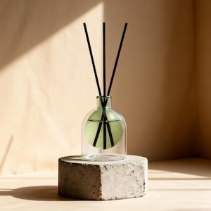 Può includere: Un diffusore a bastoncini in vetro trasparente con una camera interna sferica contenente liquido verde chiaro. Tre sottili bastoncini neri sono inseriti nel collo stretto della bottiglia e si estendono verso l'alto. Il diffusore poggia su una base in cemento grigio chiaro testurizzato.