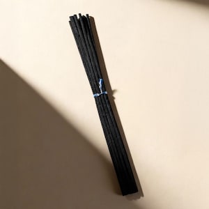 Könnte beinhalten: Ein Bündel schwarzer Räucherstäbchen, gebunden mit einem blauen Faden, vor einem beigefarbenen Hintergrund. Die Räucherstäbchen sind parallel angeordnet. Die Stäbchen haben eine strukturierte Oberfläche und sind diagonal ausgerichtet.