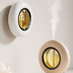 May include: Two circular humidifiers, one white and one beige, each featuring a central gold-toned reflective window and a black circular frame. Both devices emit a vertical stream of white mist from the top, with small green indicator lights visible.
