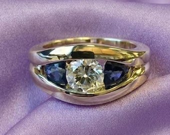 May include: A silver ring with a large, clear, round center stone flanked by two smaller, dark blue stones. The ring sits on a purple satin surface, showcasing its elegant design and sparkle. The band has a unique, curved shape.