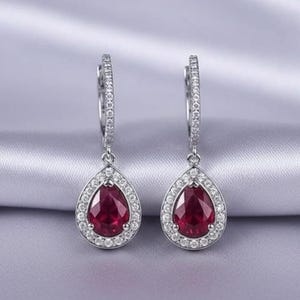 May include: Silver earrings featuring a teardrop-shaped, deep red gemstone centre, encircled by a halo of small, clear stones. The earrings have a hinged closure and are set against a soft, silvery background.