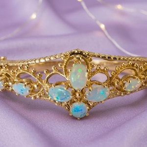May include: A gold hinged bangle bracelet featuring an intricate filigree design with small beaded accents. The bracelet is set with seven oval-shaped cabochon opals that display iridescent flashes of blue, green, and yellow light throughout.