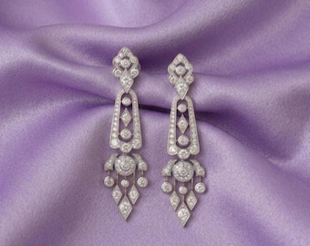 May include: A pair of silver-toned drop earrings featuring an intricate geometric design. Each earring is encrusted with numerous small, clear, round-cut stones arranged in a chandelier pattern with diamond-shaped and circular metal frames.