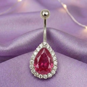 May include: A silver belly button ring with a pear-shaped, ruby-red gemstone. The gemstone is surrounded by a halo of small, clear crystals. The ring is displayed on a purple satin fabric.