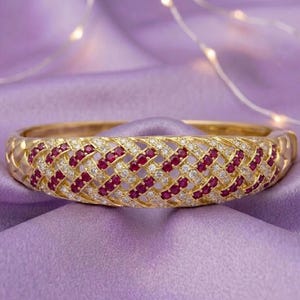 May include: A gold hinged bangle bracelet featuring a woven lattice design. The surface is encrusted with alternating rows of small, round, deep red rubies and clear, sparkling white diamonds set into the polished yellow gold metal structure.