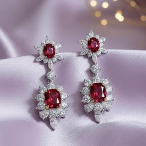May include: A pair of elegant drop earrings featuring large, square-cut, ruby-red gemstones surrounded by a halo of smaller, clear, round-cut stones. The earrings have a floral design with a silver-colored metal setting.
