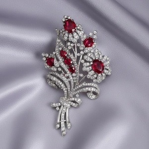 May include: A silver-toned brooch in the shape of a floral bouquet. The brooch features red gemstones at the center of the flowers and along the stems, surrounded by clear, sparkling stones. The brooch is set against a gray satin background.
