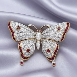 May include: A butterfly brooch with a gold frame, encrusted with sparkling white diamonds and accented with vibrant red gemstones. The wings are detailed with rows of diamonds, and the edges are lined with red stones.