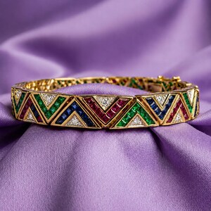 Op de afbeelding: Een scharnierende gouden armband met een herhalend geometrisch chevronpatroon. Het ontwerp is ingelegd met vierkant geslepen groene smaragden, blauwe saffieren en rode robijnen, afgewisseld met kleine, heldere, rond geslepen diamanten in driehoekige gouden zettingen.