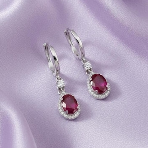 May include: A pair of silver-toned drop earrings with oval, deep red gemstones. Each earring features a halo of small, clear stones around the red gem and a smaller clear stone at the top. The earrings are set against a lavender satin background.