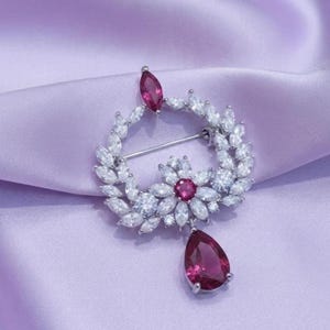 Könnte beinhalten: Eine kreisförmige Brosche mit einem Kranz aus klaren Kristallen im Marquise-Schliff, akzentuiert durch eine zentrale Blume mit einem dunkelrosa Stein und einem passenden rosa Marquise-Stein oben. Ein großer, birnenförmiger rosa Kristall hängt unten.
