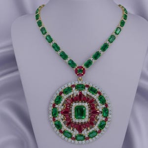 May include: A gold necklace with a large pendant. The pendant features a central emerald-cut green stone, surrounded by red and green stones, and a ring of small white stones. The necklace chain is decorated with green stones.