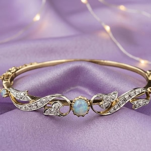 May include: A gold hinged bangle bracelet featuring a central oval opal, smaller opals at the ends, and intricate silver-toned scrollwork accents set with small clear stones. The bracelet includes a delicate safety chain attached to one side.