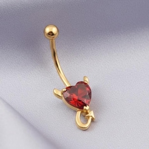 May include: A gold-toned navel ring featuring a curved barbell with a spherical top bead and a bottom charm shaped like a heart with two small horns and a pointed tail, set with a faceted red gemstone in the center of the heart design.