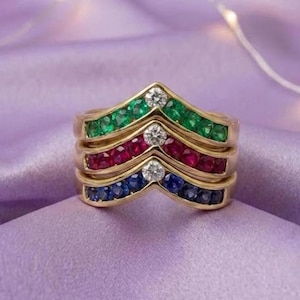 Op de afbeelding: Een gouden chevronring bestaande uit drie gestapelde banden. De bovenste band bevat groene edelstenen, de middelste band rode edelstenen en de onderste band blauwe edelstenen. Elke band heeft een enkele heldere diamant op het middelpunt.