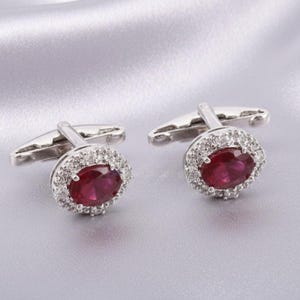 May include: Two silver cufflinks, each featuring an oval, deep red gemstone encircled by a halo of smaller, clear stones. The cufflinks are displayed on a soft, silvery background, accentuating their refined design.