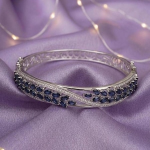 Puede incluir: Un brazalete rígido articulado de tono plateado con diseño cruzado. La pulsera está adornada con piedras azul oscuro de talla marquesa dispuestas en patrones de hojas, acentuadas por filas de pequeñas piedras claras en pavé.