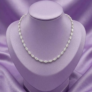 May include: A silver-toned necklace featuring a continuous chain of marquise-cut clear stones. The stones are set in a linked metal frame, creating a uniform, shimmering design that encircles the neck. The piece is displayed on a smooth form.