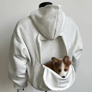 May include: A light gray hoodie with a kangaroo pouch designed to carry a small dog. The pouch is white and has a small opening. A brown and white dog is nestled inside the pouch.