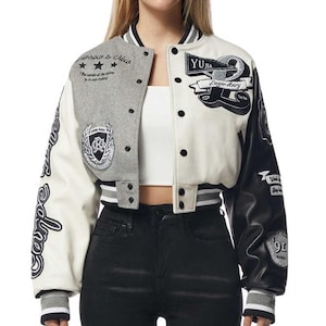 May include: Cropped varsity jacket in cream, gray, and black. The jacket features the text "Tomorrow is Now" and "Legendary" with patches and embroidered details. Ribbed cuffs and hem with black and white stripes complete the look.