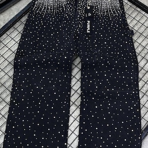 May include: Black denim jeans embellished with scattered rhinestones. The jeans feature a wide-leg design, front pockets, and the word "PURPLE" printed on a tag. The rhinestones are concentrated at the top of the jeans.