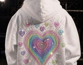 May include: A white hooded sweatshirt featuring a large, central heart design on the back, constructed from multicolored rhinestones in a rainbow gradient pattern. Smaller individual rhinestone hearts surround the central motif, with radiating lines of crystals extending outward from the center.