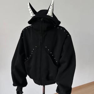 May include: Black zip-up hoodie with a hood featuring white horns with silver studs. The hoodie has silver studs along the seams and a front pocket. The sleeves end in black gloves.