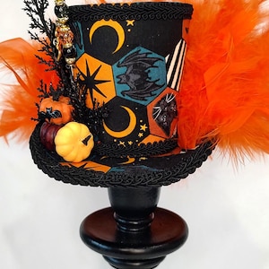 May include: A miniature top hat featuring a black fabric with orange, teal, and white geometric patterns of moons, stars, bats, and cats. It is decorated with orange feathers, small pumpkins, and black foliage, resting on a black pedestal stand.