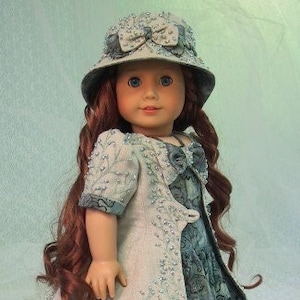 May include: A doll wearing a blue and white dress with a matching jacket and a light blue hat with a bow. The doll has long red hair and blue eyes.