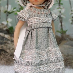 Puede incluir: Una mu&ntilde;eca de porcelana que lleva un vestido gris y blanco con ribetes de encaje y un gorro blanco. La mu&ntilde;eca tiene el pelo rojo y los ojos marrones.