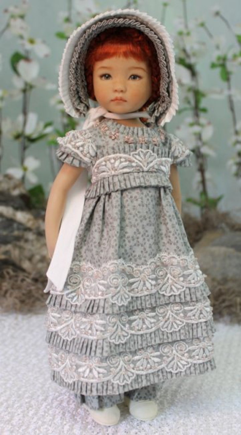 Puede incluir: Una mu&ntilde;eca de porcelana que lleva un vestido gris y blanco con ribetes de encaje y un gorro blanco. La mu&ntilde;eca tiene el pelo rojo y los ojos marrones.