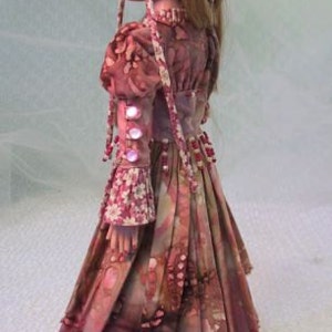 以下が含まれることがあります： 花柄のロングドレスと、お揃いのヘッドピースを身に着けたヴィンテージドール。ドレスはピンクとバーガンディの模様で、フリル袖や装飾ボタンなどのディテールが特徴です。人形は長い、明るい茶色の髪をしています。