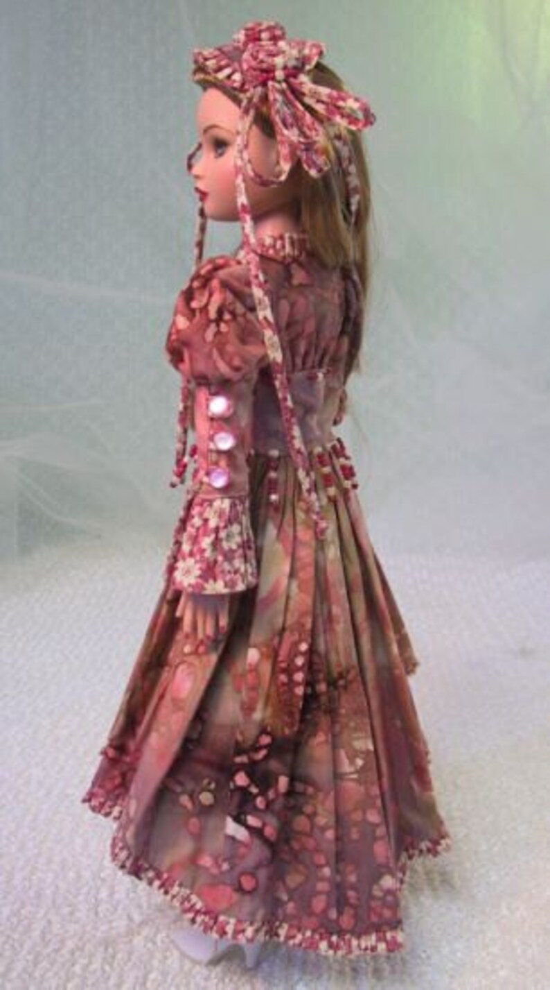以下が含まれることがあります： 花柄のロングドレスと、お揃いのヘッドピースを身に着けたヴィンテージドール。ドレスはピンクとバーガンディの模様で、フリル袖や装飾ボタンなどのディテールが特徴です。人形は長い、明るい茶色の髪をしています。