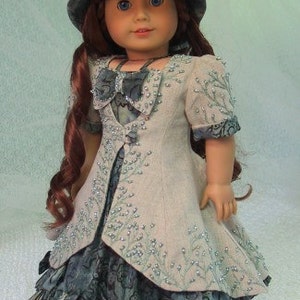 May include: A doll wearing a light gray dress with a floral pattern and a light blue hat with a bow. The doll has long red hair and blue eyes.